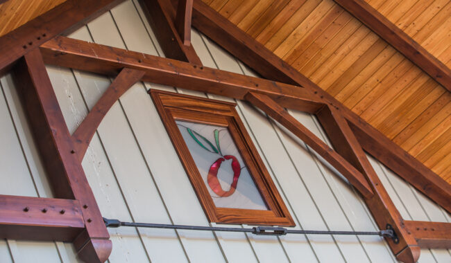 A small window with a wooden frame set in a peaked roof with exposed beams. The window features stained glass art of a red apple with a green leaf. The siding is light-colored vertical panels.
