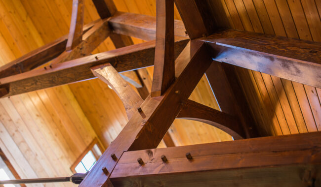 Close-up view of exposed wooden beams and joinery in a timber-framed ceiling, showcasing the warm-toned wood and craftsmanship, with natural light filtering through nearby windows.
