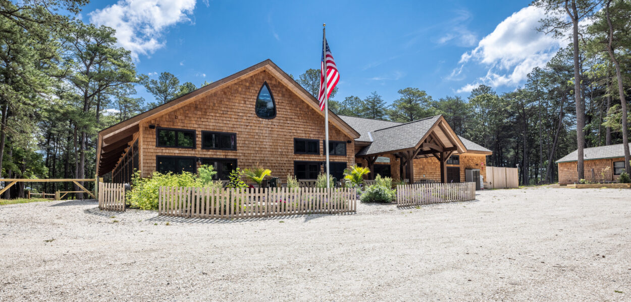 A rustic wooden building with a pointed window and a covered entrance sits among tall trees. An American flag is displayed on a pole, and a small fenced garden is in front of the building under a bright blue sky.