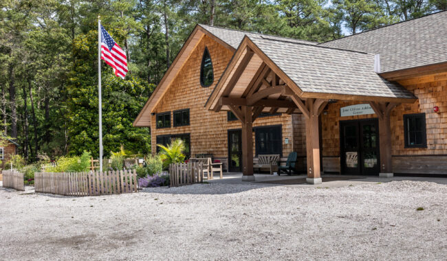 A wooden building with a gabled roof stands by a gravel parking area, with benches and potted plants near the entrance. An American flag flies on a tall pole in front, surrounded by trees and a small fenced garden.