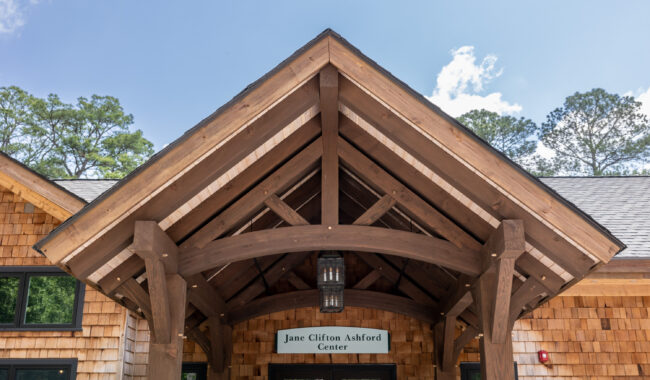 A wooden entryway with exposed beams and a hanging lantern leads to a building with shingle siding. A sign above the door reads “Jane Clifton Ashford Center.” Trees and a blue sky are visible in the background.