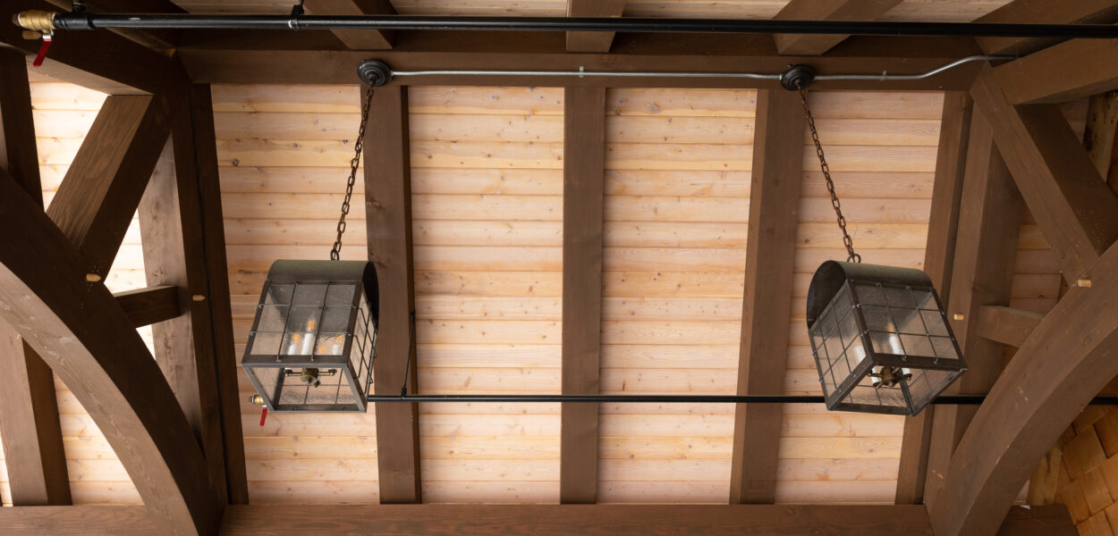 Two black metal lanterns hang from chains on a rectangular metal bar attached to a wooden ceiling with exposed beams and light-colored wood planks.