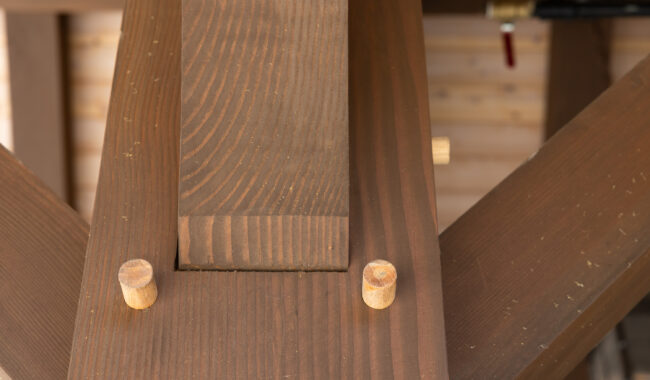 Close-up of a wooden joint in timber framing, showing interlocking beams secured with round wooden pegs. The wood has a smooth finish and visible grain patterns. A blurred valve is in the background.