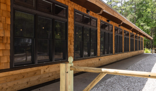 A long wooden building with large black-framed windows, shingle siding, a sloped roof, and a wooden fence in front, surrounded by trees and sunlight.