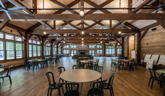A spacious wooden hall with high ceilings, exposed beams, large windows, and multiple round tables with chairs. The room has polished wood floors, ceiling fans, and natural light streaming in.