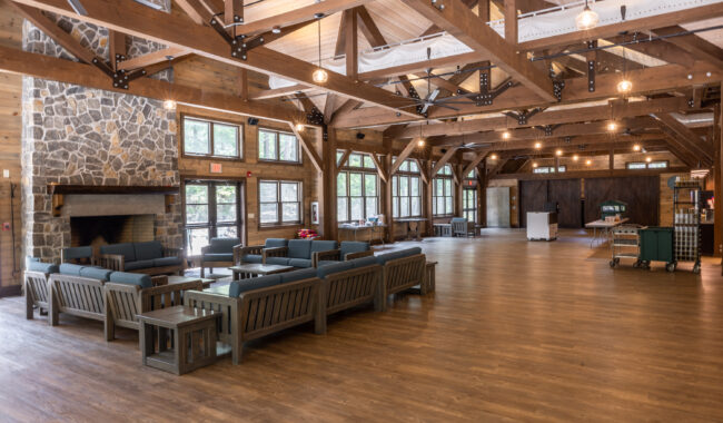 Spacious lodge interior with wooden beams, large stone fireplace, and windows letting in natural light. Blue cushioned seating is arranged in the center on a wooden floor. The space appears open, bright, and inviting.