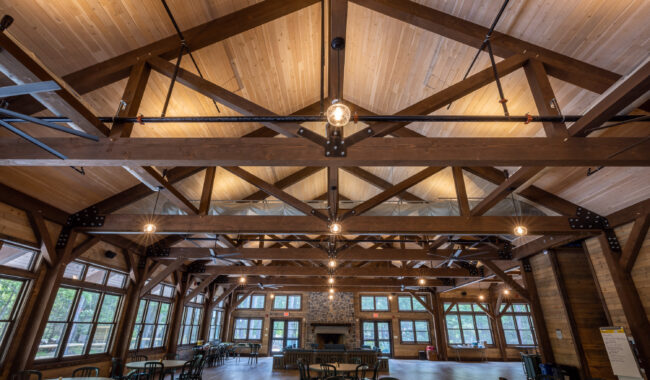 Spacious wooden hall with exposed beams, large windows, and high ceiling. Round tables and chairs are arranged throughout. A stone fireplace is centered on the far wall, and hanging lights illuminate the room.