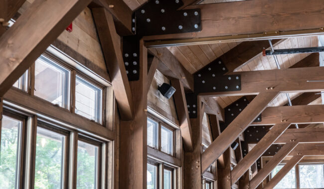 Exposed wooden beams and black metal brackets support a high ceiling with large windows along the wall, letting in natural light and offering views of trees outside.