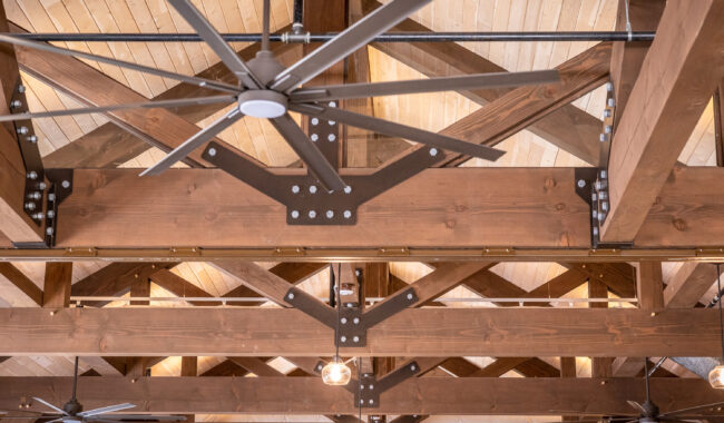 Large metal ceiling fan attached to exposed wooden beams and trusses in a high, vaulted ceiling with light wood paneling. Additional fans and hanging lights are visible in the background.