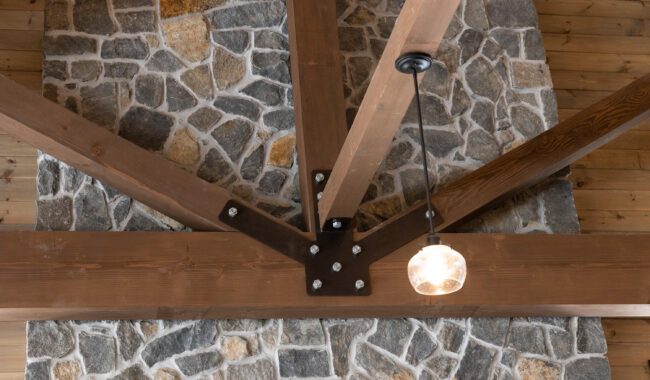 Close-up view of a wooden ceiling with exposed beams, metal brackets, and bolts, intersecting above a stone wall. A single glass pendant light hangs from the ceiling near the beams.