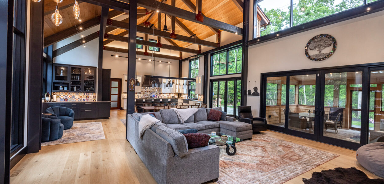 Modern open-concept living room with high wood-beamed ceilings, large windows, gray sectional sofa, rugs, and a spacious kitchen in the background. Natural light fills the space, with views of trees outside.