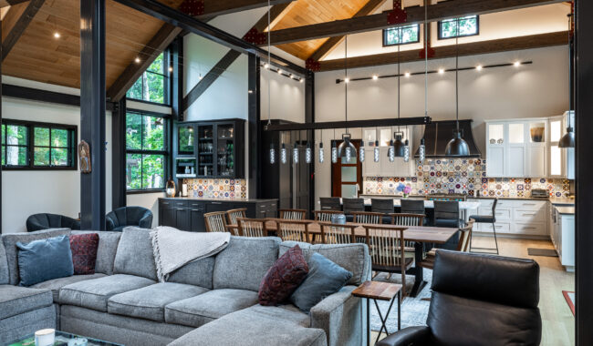 Spacious modern open-concept living room and kitchen with exposed wooden beams, large windows, a gray sectional sofa, dining table, pendant lights, and colorful tiled backsplash in the kitchen.