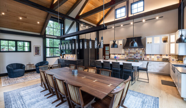 Spacious modern kitchen and dining area with high vaulted wooden ceilings, large windows, a long wooden dining table, black chairs, pendant lighting, and colorful tile backsplash.