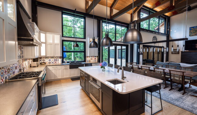 Spacious modern kitchen with large windows, high wood-beamed ceilings, a central island with a sink, pendant lights, white cabinets, patterned backsplash, and an open dining area with a wooden table and chairs.