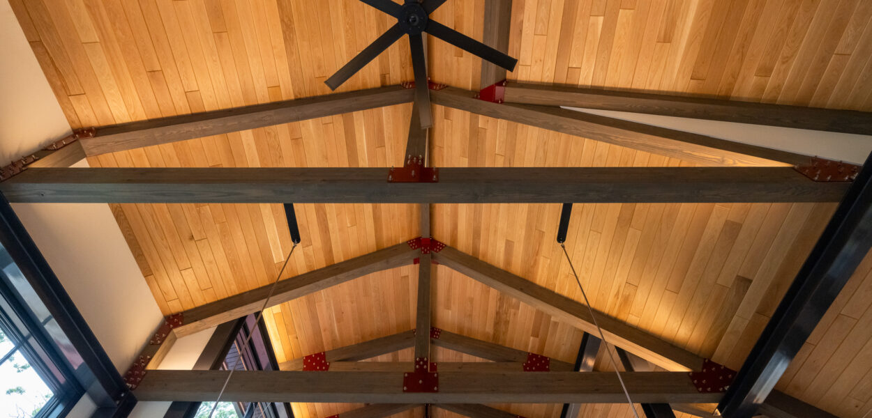 Ceiling view of a modern room with exposed wooden beams, a large black ceiling fan, wood paneling, and metal hardware joining the beams. Natural light is visible from windows at the edges.