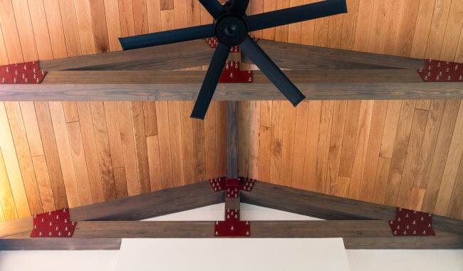 A black ceiling fan hangs from a wooden vaulted ceiling with exposed dark wooden beams and red metal brackets at the joints. The ceiling panels are a light natural wood.