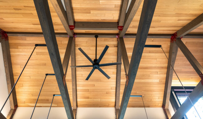 A wooden ceiling with exposed beams and a central black ceiling fan. The beams are reinforced with red metal brackets and black cables, and natural light enters from a window on the right.