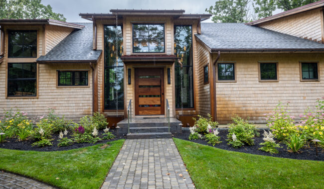 Modern house with tan shingle siding, large windows, and a wooden front door with horizontal glass panels. A stone pathway leads to the entrance, surrounded by green lawn and landscaped flower beds.