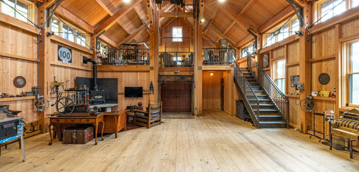 Spacious wooden workshop with high vaulted ceiling, exposed beams, large fans, metal staircase, vintage tools, machinery, and loft area. Bright natural light streams through multiple windows.