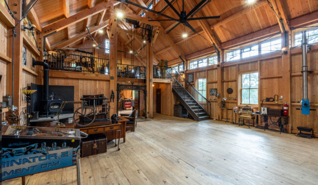 Spacious, rustic wooden interior with high ceilings, exposed beams, large windows, and vintage machinery. A metal staircase leads to a loft area, and various antique tools and equipment line the walls.
