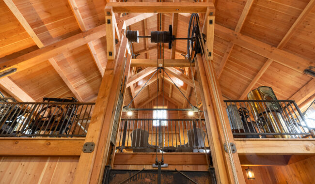 Interior view of a wooden building with exposed beams and vintage industrial machinery, including pulleys and wheels, displayed on a loft with metal railings. Sunlight streams in from windows above.