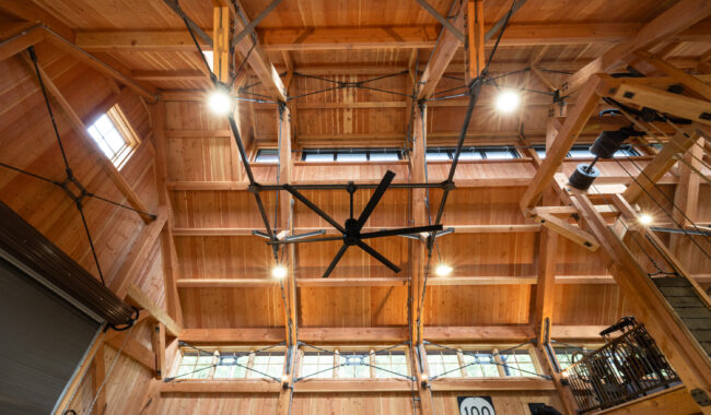 View of a high wooden ceiling with exposed beams, metal supports, and a large industrial ceiling fan in a spacious, well-lit interior, possibly a barn or warehouse. Bright lights and windows provide natural light.