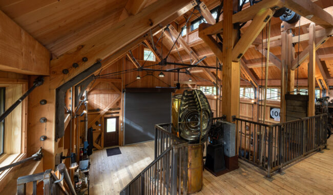 A spacious loft with exposed wooden beams, industrial metal railings, large mechanical gears, and tall windows letting in natural light. A door and a roll-up garage door are visible on the lower level.