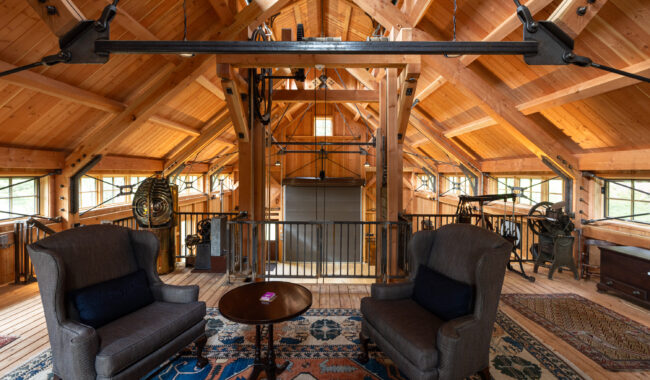 A cozy loft with exposed wooden beams, two armchairs around a round table, patterned rugs, large windows, and visible mechanical equipment and gears along the walls.