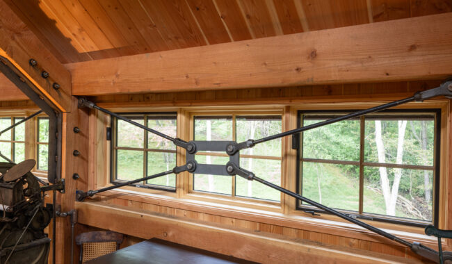 Interior view of a wooden room with large horizontal windows, showcasing metal cross-bracing beams attached to the walls and ceiling. Trees and greenery are visible outside through the windows.