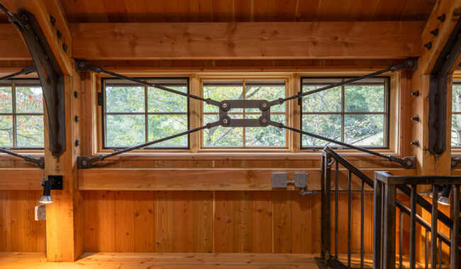 Interior view of a wooden loft with large horizontal windows framed by exposed metal structural supports, letting in natural light and showing trees outside. A metal railing is visible on the right side of the image.