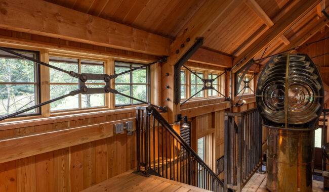 A wooden interior with large windows, exposed beams, metal supports, and a lighthouse Fresnel lens on a brass base, overlooking a staircase and lush greenery outside.