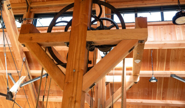 A close-up of a large pulley system with wooden beams and metal wheels mounted indoors, under a ceiling with exposed wood and windows letting in natural light.