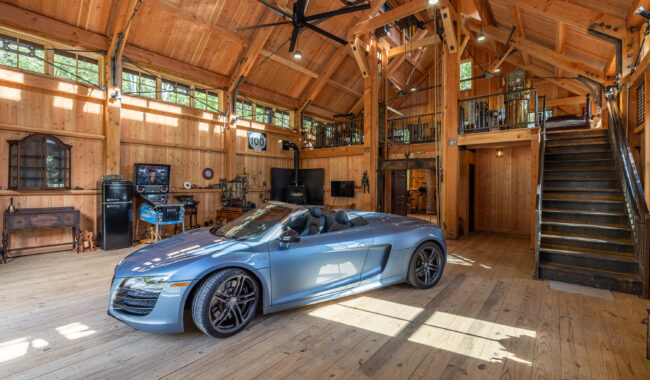 A silver convertible sports car is parked inside a spacious, rustic wooden garage with high ceilings, large windows, and a metal staircase leading to a loft area. Various tools and equipment are visible around the space.