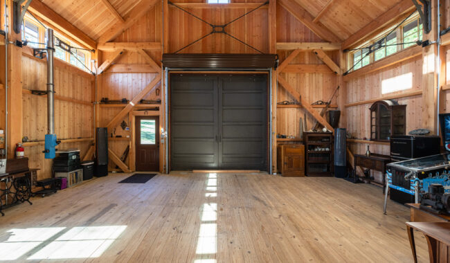 Spacious wooden barn interior with high ceilings, exposed beams, large black double doors, several windows letting in natural light, and vintage furniture along the walls.
