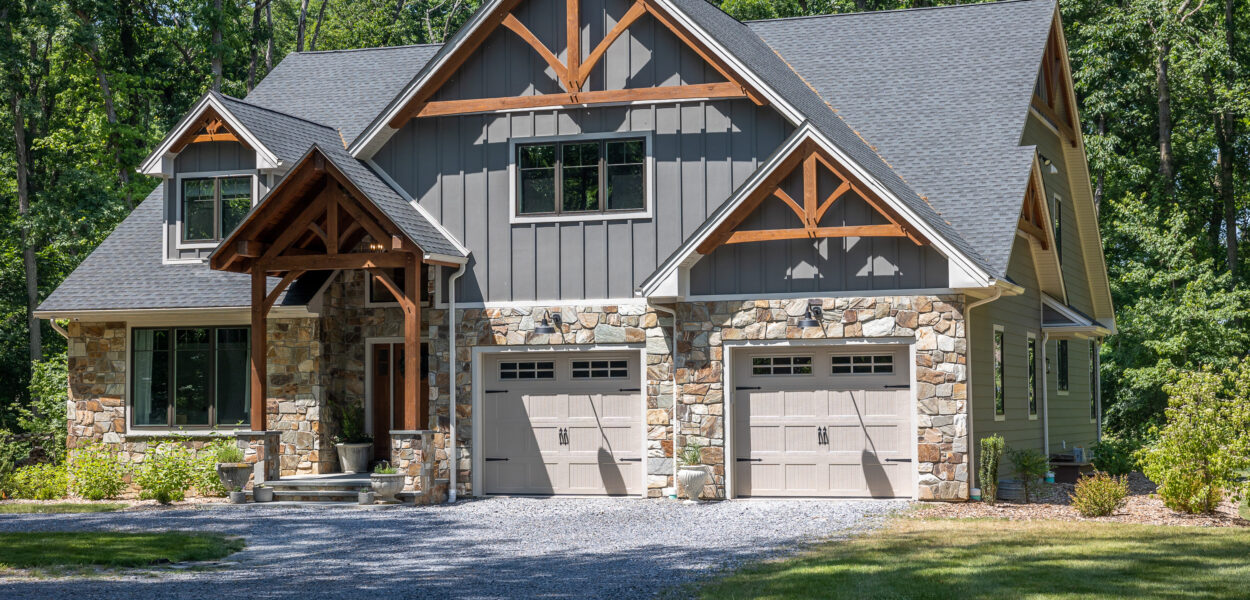 A modern craftsman-style house with a stone and wood exterior, two garage doors, a covered front porch, and surrounded by trees and greenery.