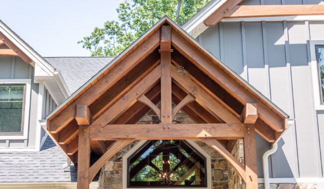 A close-up view of a wooden gabled porch roof with exposed beams and a stone wall, attached to a modern gray house with board and batten siding. A bird’s nest is visible in the beams.