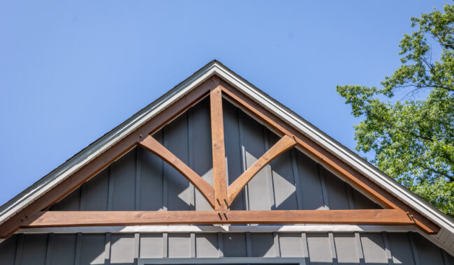 Gable roof of a modern house with gray vertical siding and exposed decorative wooden trusses, set against a clear blue sky with green tree branches visible on the right side.