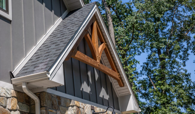 Close-up of a house exterior showing a peaked roof with gray shingles, stone walls, board and batten siding, and a decorative wooden truss above a small gable, with trees and blue sky in the background.