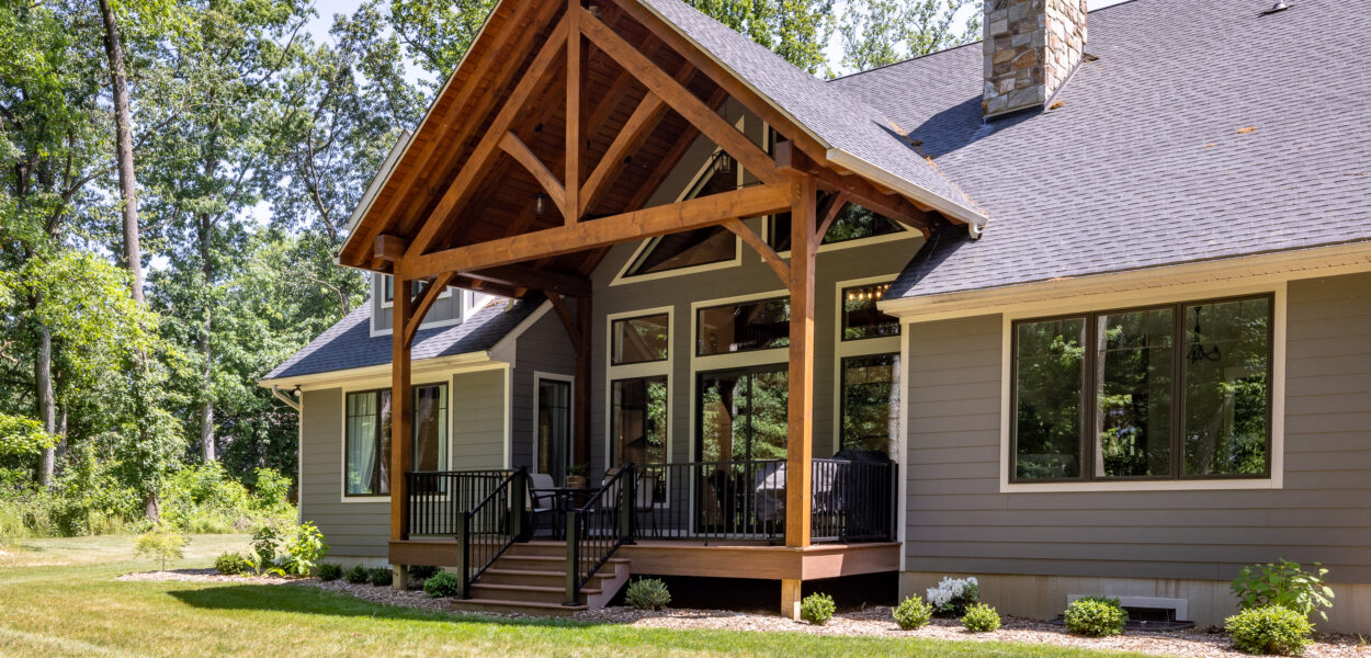 A modern house with gray siding, large windows, and a peaked wooden porch with exposed beams, set in a grassy yard surrounded by tall trees on a sunny day.
