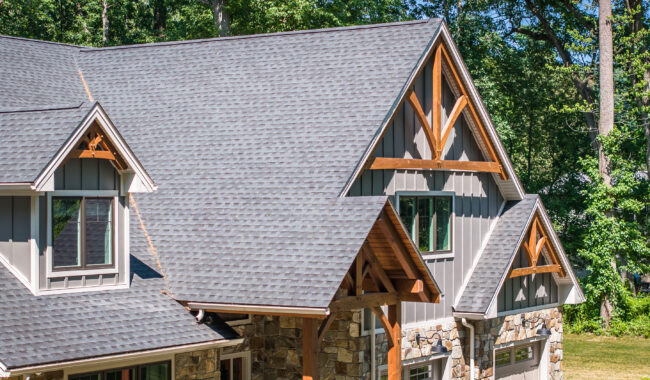 A close-up view of a modern house with gray shingles, wooden beams, stone accents, and gray siding, surrounded by green trees on a sunny day.