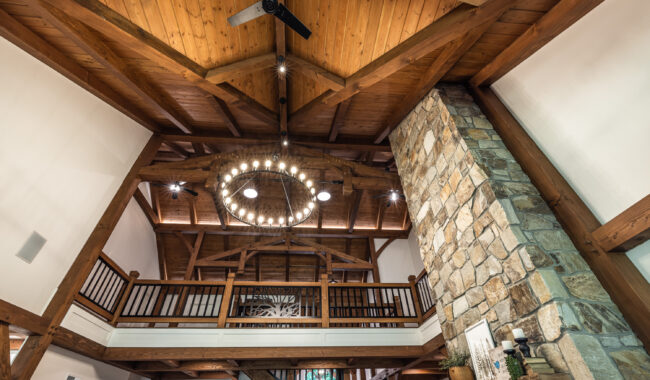 Looking up at a rustic interior with exposed wooden beams, a circular chandelier, a stone fireplace, and a wooden balcony railing on the second floor, all illuminated by warm lighting.