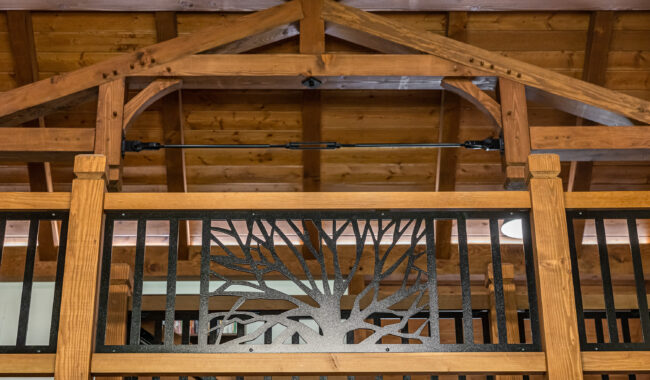 Wooden balcony railing with a metal design of a tree, viewed indoors. The background features exposed wooden beams and a high, peaked ceiling.