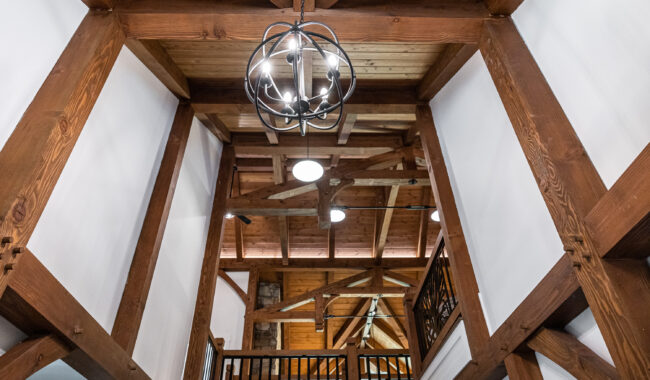View of a timber frame ceiling with exposed wooden beams, white walls, a modern metal chandelier, and a loft railing, showcasing a rustic, open architectural design.
