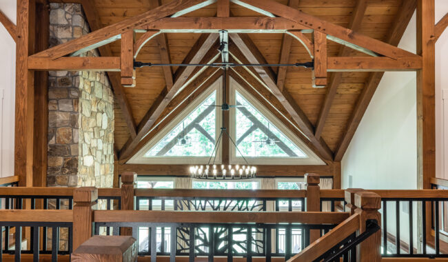 A loft area with exposed wooden beams, a large triangular window with geometric panes, a stone chimney, black railings, and a modern chandelier hanging from the ceiling.