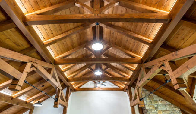 Exposed wooden beam ceiling with triangular trusses, warm lighting, and a skylight above a white wall with a door and part of a stone accent visible on the right.