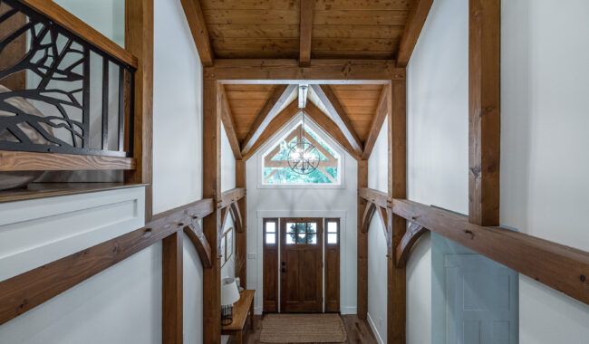 A bright entryway with wooden beams, vaulted ceiling, a large wooden front door with glass panels, sidelights, and a woven rug on hardwood floors. A geometric chandelier hangs from the ceiling.