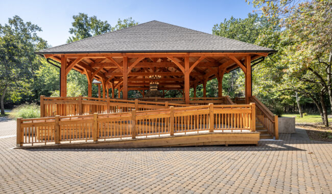 A wooden pavilion with a gray roof stands in a park setting. A wide, accessible wooden ramp curves up to the entrance, surrounded by trees and a paved walkway under a clear blue sky.