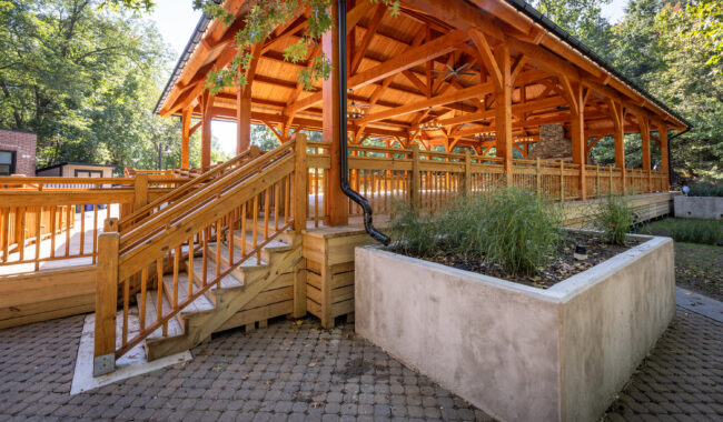 A large wooden pavilion with exposed beams and a gabled roof stands in a park. A ramp and stairs with wooden railings lead up to it, and greenery is planted in a raised concrete bed nearby. Trees surround the structure.