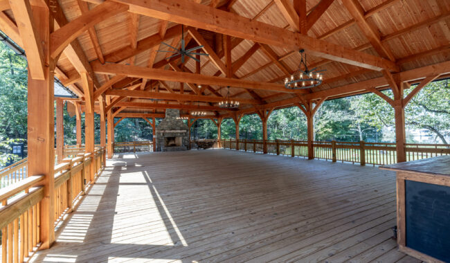 Large open wooden pavilion with exposed beams, a high peaked roof, chandeliers, and a stone fireplace at one end. The space is surrounded by a wooden railing and overlooks green trees and grass outside.