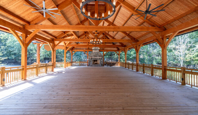 A spacious, open wooden pavilion with a high vaulted ceiling, large chandeliers, ceiling fans, and a stone fireplace at the far end. Sunlight streams in through the open sides, revealing trees outside.